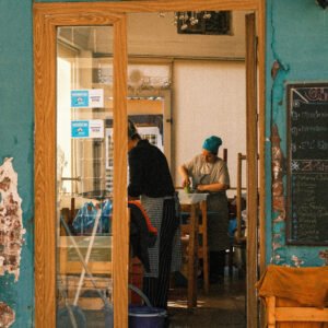 A photograph of a woman preparing vegetables in a restaurant, seen through a doorway, the colours are teal, turquoise, and woody. It is reminiscent of Vermeer