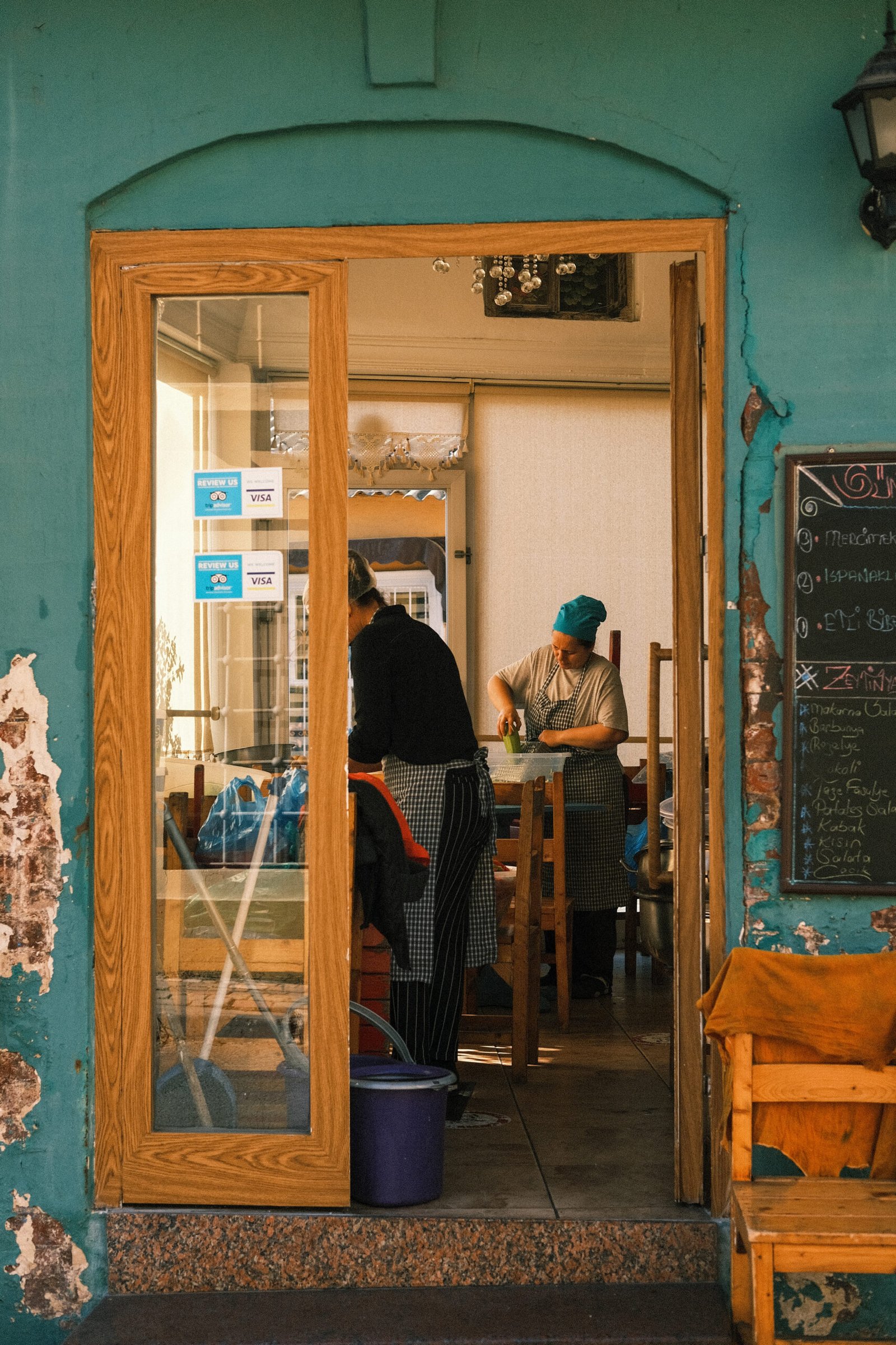A photograph of a woman preparing vegetables in a restaurant, seen through a doorway, the colours are teal, turquoise, and woody. It is reminiscent of Vermeer