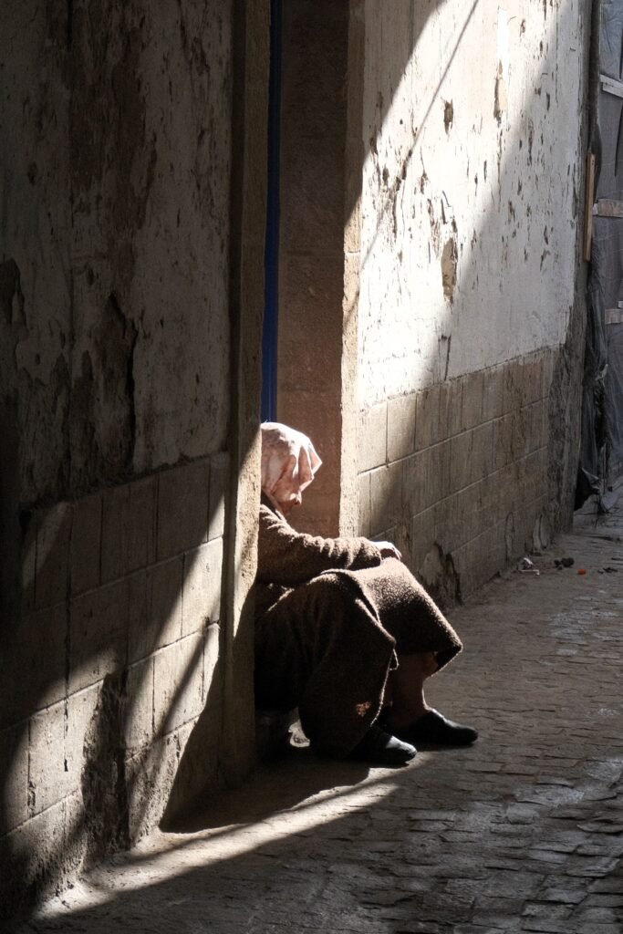 Morning light shining down on an elderly veiled Moroccan woman sitting and begging in a doorway. Cobbled streets, flaking white walls