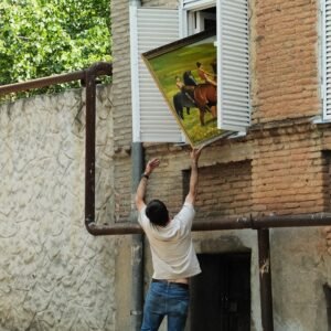 A photograph of a man on tiptoes in an unintentionally balletic pose pushing a framed oil painting through the window of a Tbilisi townhouse.